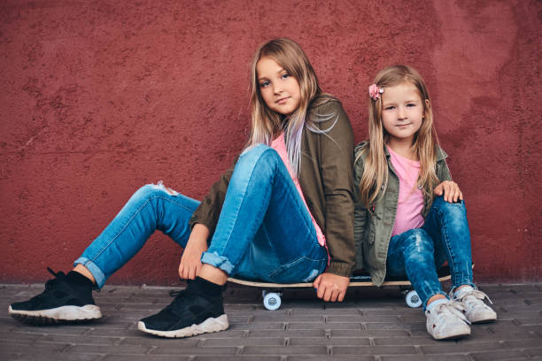 Portrait of two little sisters dressed in trendy clothes leaning on a wall while sitting on a skateboard at bridge footway.