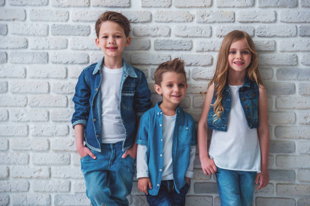 Cute little kids in stylish jeans clothes are looking at camera and smiling, standing against white brick wall